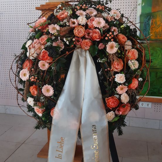 Ein Blumenkranz aus verschiedenen Blumen wie Rosen, Gänseblümchen und Grünpflanzen mit einem weißen Band, auf dem 'In Liebe' steht. Das Band ist an einem Holzständer befestigt und auf einem gefliesten Boden platziert.