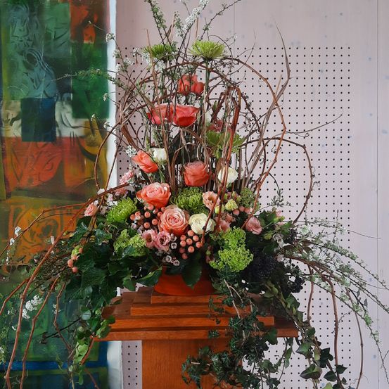 Ein hölzernes Podium präsentiert eine aufwendige Blumenarrangement mit einer Mischung aus roten, rosa und weißen Blumen, ergänzt durch grüne Blätter und Zweige, vor einer weißen Wand mit einem Muster aus Löchern.