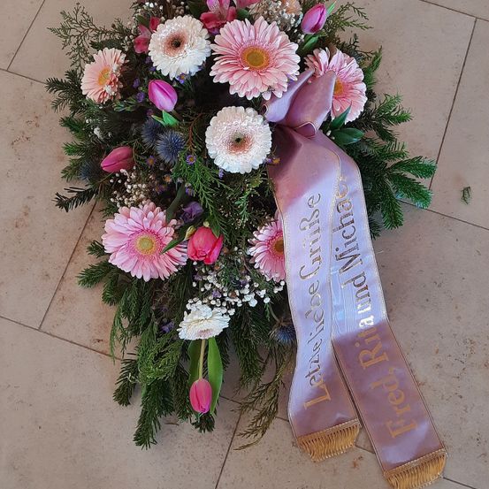 Ein Trauerkranz mit rosa und weißen Blumen, einer Schleife mit den Namen Fred und Michael und Grünpflanzen auf einem gefliesten Boden.