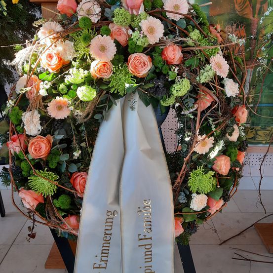 Ein Kranz mit rosa und weißen Blumen, grünen Blättern und einer Schleife mit goldener Schrift. Die Schleife trägt die Aufschrift 'Erinnerung' und 'Frühling und Familie.' Der Kranz steht auf einer weißen Oberfläche, möglicherweise einem Tisch.