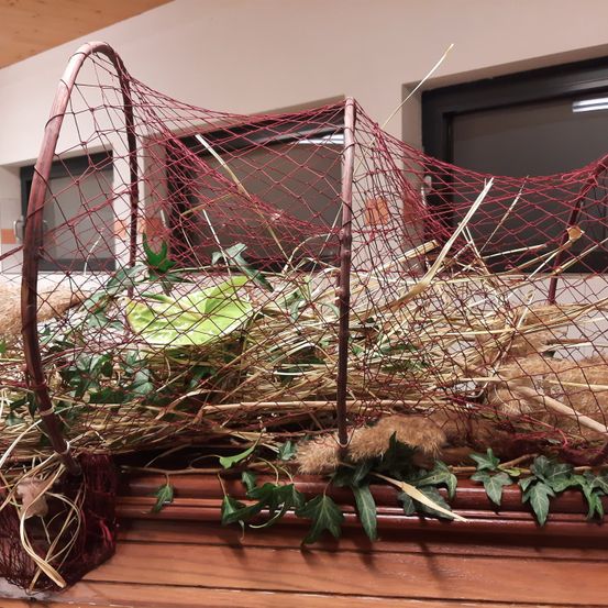 Ein dekoratives rotes Netz ist auf einem Holzsims angeordnet, gefüllt mit Stroh und grünen Blättern. Das Netz hat eine gebogene Form und steht vor einem Fenster.