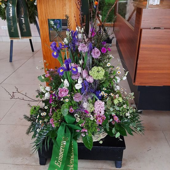 Ein farbenfrohes Blumenarrangement mit lila Iris, rosa Rosen und weißen Gänseblümchen steht in einem Korb auf einem schwarzen Ständer, begleitet von grünen Bändern. Das Bouquet befindet sich auf einem gefliesten Boden, mit einer hölzernen Struktur im Hintergrund.