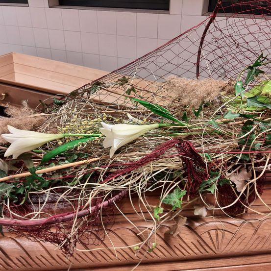 Ein hölzerner Sarg, geschmückt mit weißen Lilien, rotem Netz und getrocknetem Gras, ruht auf einer Holzfläche mit einer gefliesten Wand im Hintergrund.