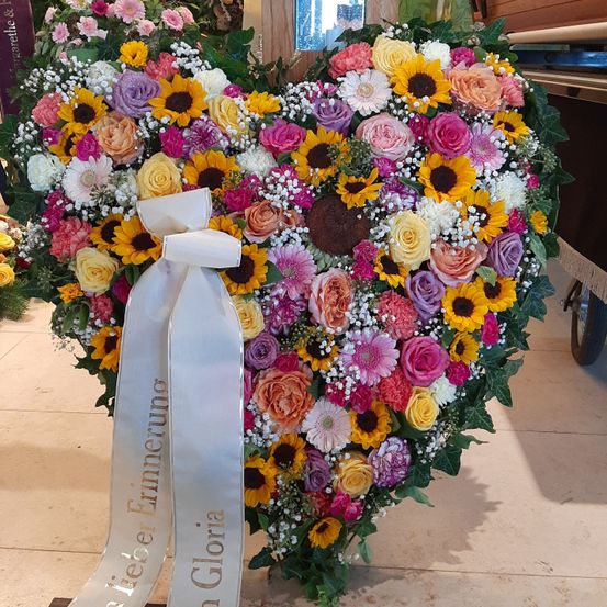 Ein herzförmiges Blumenarrangement mit einer weißen Schleife, die 'A Teier Erinnerung in Gloria' besagt, gefüllt mit mehrfarbigen Blumen, einschließlich Rosen, Sonnenblumen und Babys Atmung, auf einem gefliesten Boden.