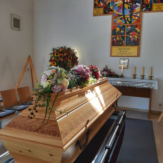 Ein hölzerner Sarg, geschmückt mit Blumen, steht in einer Kapelle. Dahinter zeigt eine Wand ein farbenfrohes Kunstwerk. Rechts neben dem Sarg steht ein Tisch mit drei Kerzen. Links sind Stühle aufgestellt.