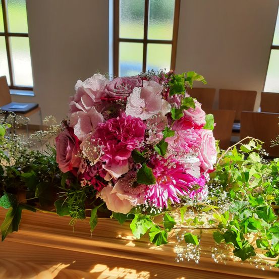 Ein großer Strauß pinker Blumen steht auf einem Holzsarg in einem Raum, durch dessen Fenster Sonnenlicht scheint. Grünpflanzen und Blätter sind um den Sarg verstreut.
