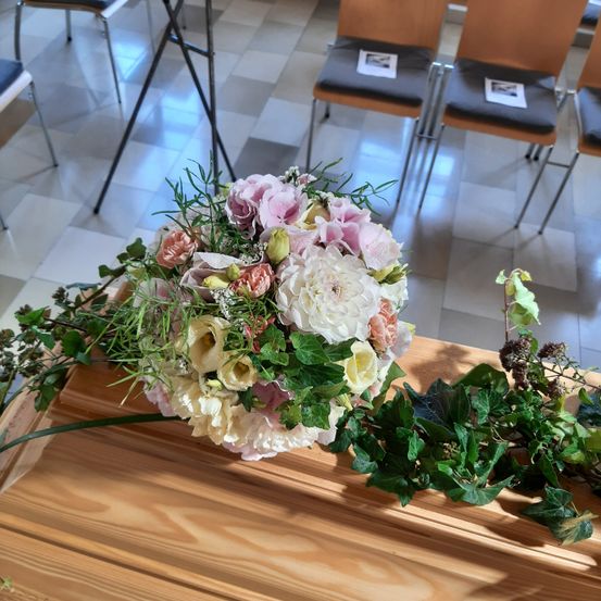 Ein großer Blumenstrauß mit rosa, weißen und gelben Blumen ruht auf einem Holzsarg. Stühle mit grauen Kissen sind im Hintergrund aufgestellt.