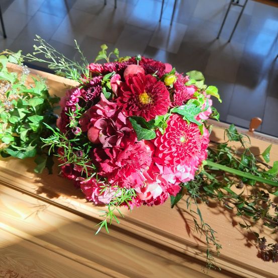Ein leuchtender Strauß roter Dahlien mit grünen Blättern liegt auf einer Holzoberfläche, möglicherweise einem Sarg, in einem gut beleuchteten Raum.