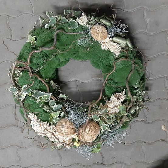 Ein Blick von oben auf einen kreisförmigen Kranz auf einem Ziegelboden. Der Kranz besteht aus grünen Blättern und Zweigen, mit drei Kokosnüssen und weißen Blumen.