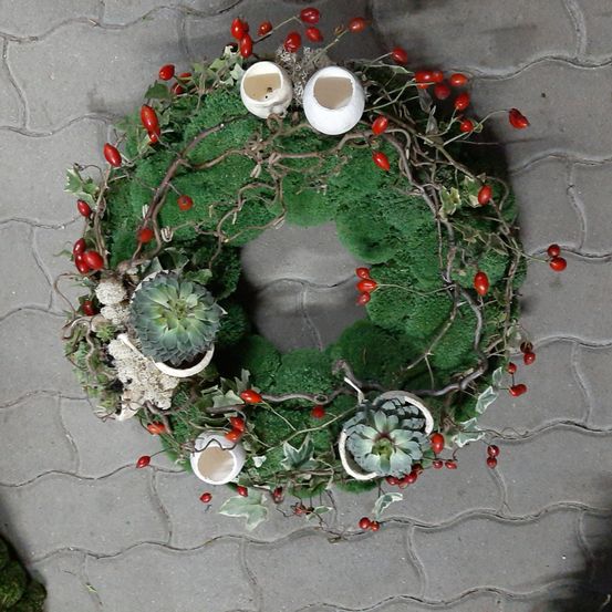 Ein Kranz aus grünem Moos und roten Beeren mit drei kleinen weißen Kerzenhaltern auf einer Betonoberfläche.