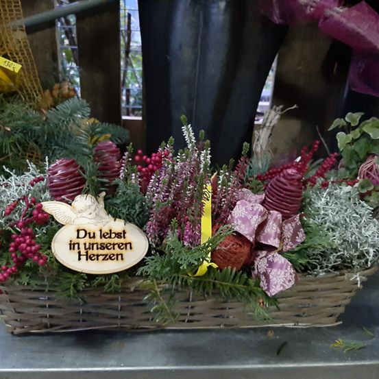 Ein Blumenkorb mit einem Schild, das 'Du lebst in unseren Herzen' besagt, mit roten Beeren, silbernen Blättern und einem lila Band. Dahinter ist ein verschwommener Hintergrund mit einem gelben Etikett.