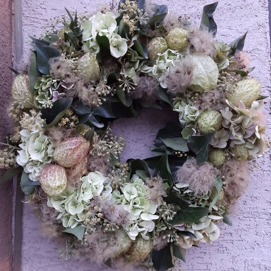 Ein Kranz aus weißen Blumen, grünen Blättern und einigen federartigen Elementen hängt an einer rosa Wand.