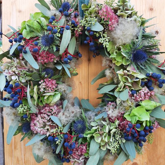 Ein farbenfroher Kranz aus verschiedenen Blumen, Blättern und Beeren, der auf einer Holzoberfläche präsentiert wird. Der Kranz zeigt Blau-, Rosa- und Grüntöne.