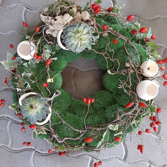 Ein Kranz aus grünem Moos mit roten Beeren und Sukkulenten. Vier weiße Kerzenhalter sind um den Kranz verstreut.