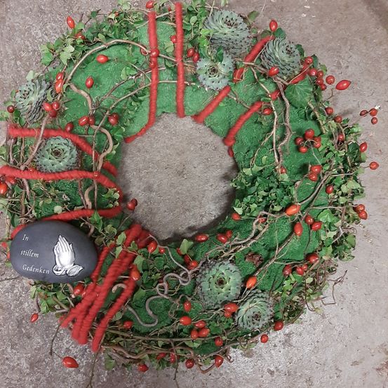 Ein Kranz aus grünem Moos, roten Beeren und Sukkulenten mit einem schwarzen Stein, der ein betendes Händedesign und die Worte 'In stillem Gedenken' zeigt, liegt auf einer grauen Oberfläche.