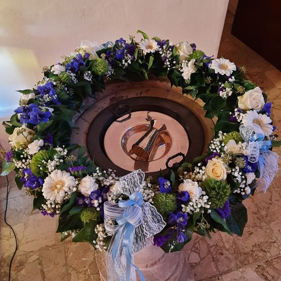 Ein runder Taufbrunnen, geschmückt mit einer Girlande aus weißen und blauen Blumen und grünen Blättern, mit einem silbernen Kelch in der Mitte. Der Hintergrund besteht aus einer weißen Wand und einem gefliesten Boden.