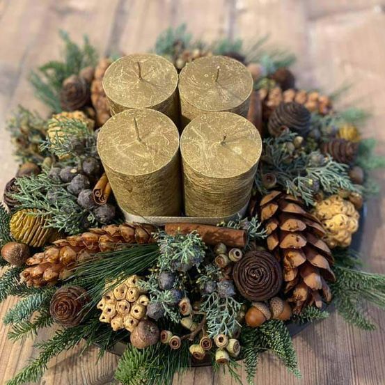 Ein hölzerner Tisch trägt einen Kranz aus Tannenzapfen, grünen Blättern und goldenen Kerzen. Der Kranz ist mit drei Kerzen in der Mitte und umgeben von verschiedenen natürlichen Elementen dekoriert.