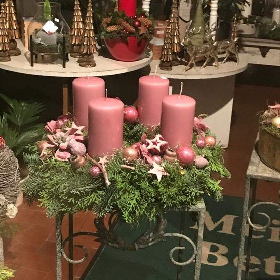 Ein Weihnachtskranz mit vier rosa Kerzen steht auf einem Metallständer. Es gibt Tannenzweige, rote Ornamente und Sterne im Kranz. Im Hintergrund stehen Tische mit Weihnachtsdekorationen.