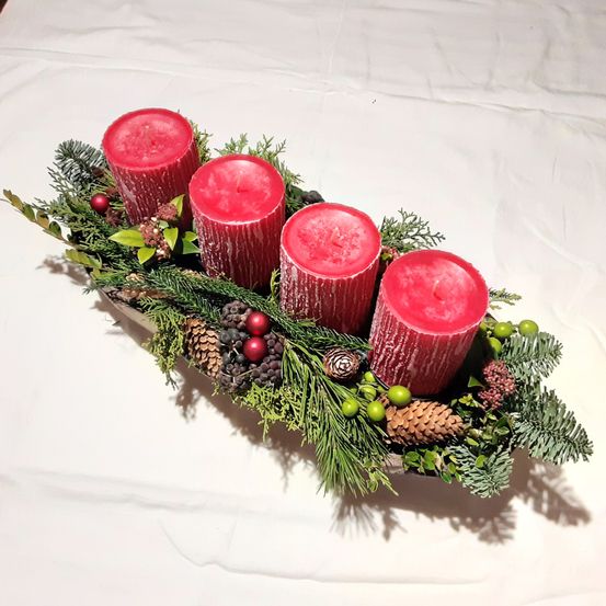Ein silbernes Tablett hält fünf rote Kerzen und ist von grünen Tannennadeln und Tannenzapfen umgeben. Die Umgebung ist auf einer weißen Oberfläche.