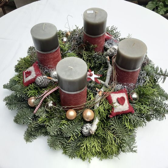Ein Kranz aus immergrünen Pflanzen hält vier Kerzen. Zwei sind hoch und zwei sind kurz. Er hat rote Ornamente, Tannenzapfen und kleine rote Kissen.
