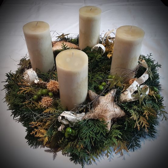 Ein Kranz mit vier weißen Kerzen, umgeben von Grün und dekorativen Elementen wie einem Stern und Bändern. Er ruht auf einer weißen Oberfläche.