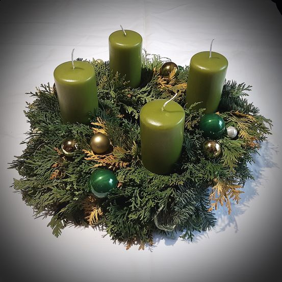 Ein Kranz mit grünem Laub und vier grünen Kerzen, verziert mit grünen und goldenen Kugeln, steht auf einer weißen Oberfläche.