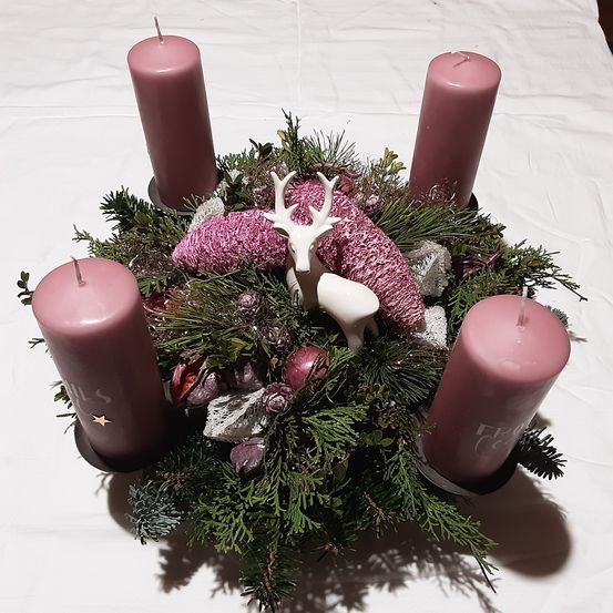 Ein Kranz mit vier rosa Kerzen, grünen Blättern und einer weißen Hirschfigur. Der Kranz liegt auf einem weißen Tuch.