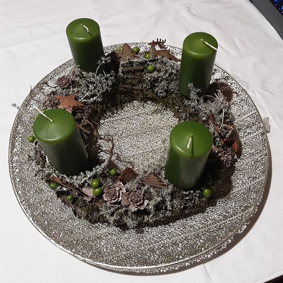 Ein silberner Teller hat einen Weihnachtskranz mit vier grünen Kerzen, grünen Blättern und getrockneten Blumen. Er steht auf einem weißen Tuch.