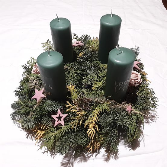 Ein Kranz mit vier grünen Kerzen und etwas Grün. Eine Kerze rechts hat das Wort 'Frohe' darauf geschrieben. Es gibt drei rosa Sterne und zwei rosa Blumen am Kranz.