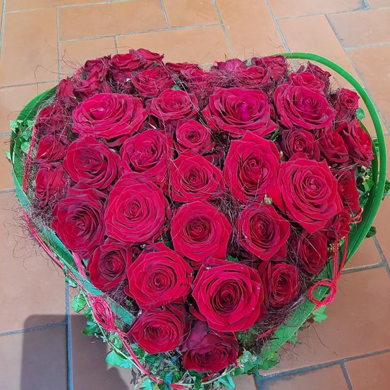 Ein herzförmiges Arrangement aus leuchtend roten Rosen in einem grünen Korb, auf einem gefliesten Boden platziert, mit einem Schatten nach links geworfen.