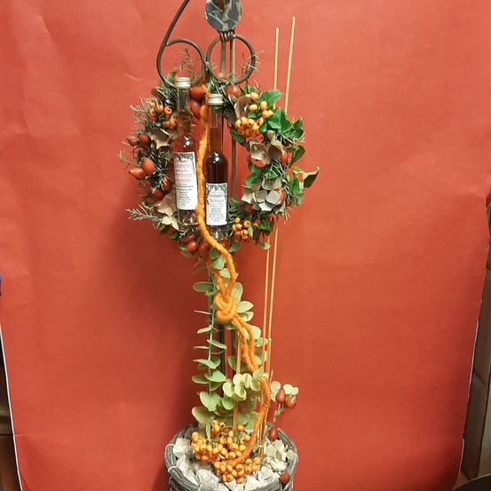 Eine dekorative Anordnung zeigt einen Kranz mit orangefarbenen Beeren und grünen Blättern, zwei Flaschen mit Flüssigkeit und einem gelben Seil vor einem roten Hintergrund.
