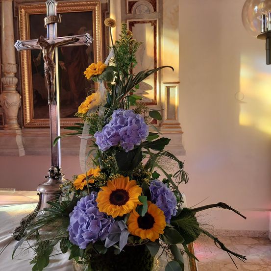 Eine Blumenarrangement aus lila und gelben Blumen in einem Vasen, mit einem Kruzifix dahinter und einem Gemälde im Hintergrund.