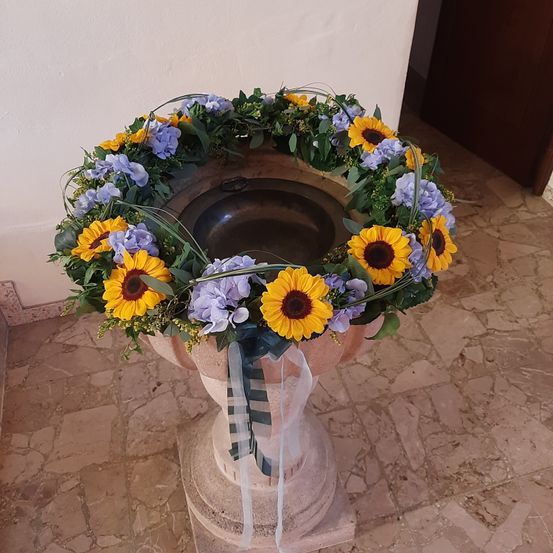 Eine dekorative Blumenarrangement aus Sonnenblumen und lila Blumen umgibt eine Taufschale. Die Schale steht auf einem Steinsockel, geschmückt mit einem Band, auf einem gefliesten Boden.
