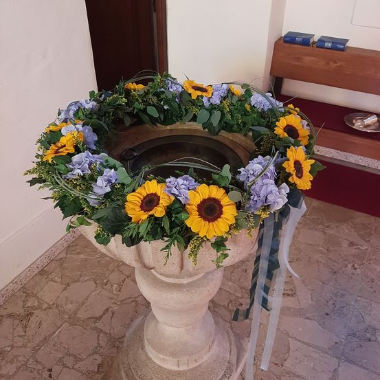 Ein Taufbecken mit Sonnenblumen und blauen Blumen steht in einer Kirche. Ein Holzbank und Bücher sind in der Nähe.