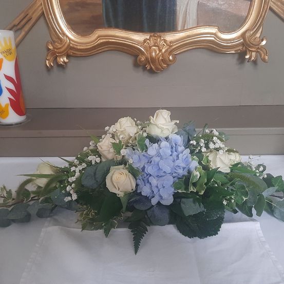 Eine weiße Tischdecke mit einer Blumenarrangement aus weißen Rosen und blauen Hortensien. Eine Kerze mit bunten Mustern und ein Spiegel mit goldenem Rahmen sind hinter den Blumen.
