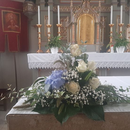 Ein Blumenarrangement steht auf einem Tisch in einer Kirche. Der Tisch ist mit einem weißen Tuch bedeckt. Dahinter steht ein goldener Altar mit Pflanzen und Kerzen. Eine rote Fahne steht links.