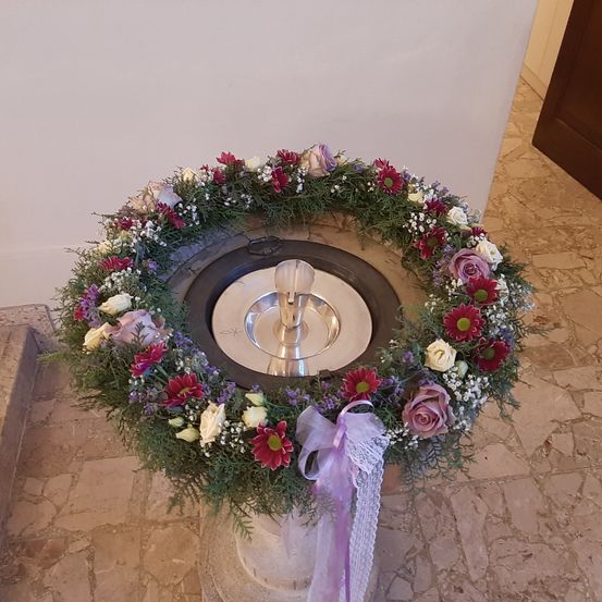 Eine Taufschale mit einem silbernen Kelch in der Mitte, umgeben von einem Kranz aus roten, weißen und lila Blumen. Der Boden besteht aus Steinfliesen.