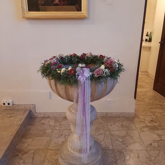 Ein großer steinerner Vasen mit einem Blumenkranz steht auf einem gefliesten Boden, verziert mit lila Bändern und einem kleinen silbernen Gegenstand. Ein gerahmtes Gemälde hängt an der Wand darüber.