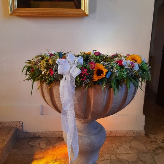 Ein großer dekorativer Steinpflanzer mit einer Vielzahl von Blumen, darunter Sonnenblumen, Gänseblümchen und Rosen, geschmückt mit einem weißen Band. Er steht auf einem gefliesten Boden.