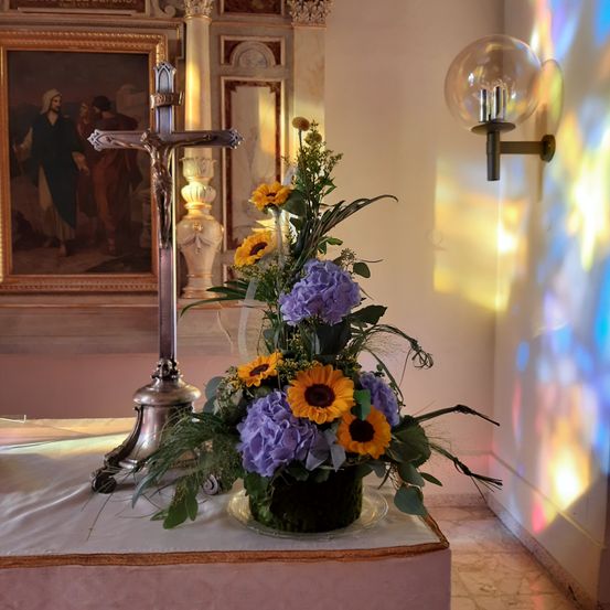 Ein Kreuz steht neben einer Blumenarrangement aus Sonnenblumen und lila Blumen auf einem Tisch in einer Kirche. Die Wand ist weiß gestrichen, und ein Buntglasfenster wirft farbiges Licht.
