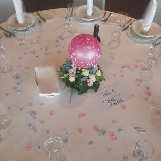 Ein Tisch ist für eine Party vorbereitet mit einem rosa Luftballon, der 'Es ist ein Mädchen' sagt, einem Blumenstrauß und einem Schild mit 'Tison S 8 Haustiere, 1 Hodswitz'. Es gibt Teller, Weingläser und Servietten auf dem Tisch.