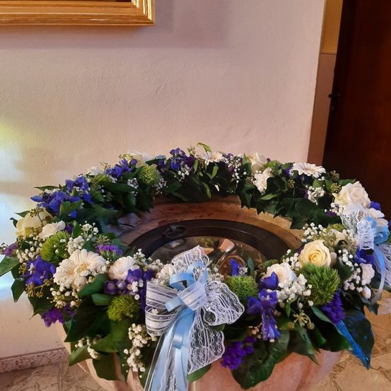 Eine Taufe mit einer kreisförmigen Blumengirlande in Blau, Weiß und Grün, mit einer blauen Schleife in der Mitte, vor einer weißen Wand mit goldenem Rahmen.