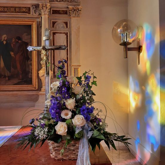 Ein Blumenarrangement auf einem Tisch mit einem Kreuz dahinter. Das Kreuz ist an einer weißen Wand montiert. Die Blumen sind in einem Korb mit einer Schleife. Ein Bilderrahmen an der Wand mit einer biblischen Szene.