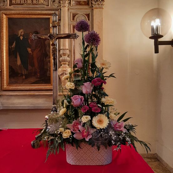 Ein großer Blumenstrauß steht auf einem roten Tisch in einer Kirche, mit einem Kreuz und einem religiösen Gemälde im Hintergrund.