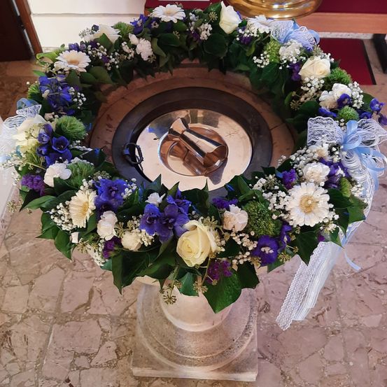 Ein Taufbecken, geschmückt mit einer Girlande aus weißen und blauen Blumen und Blättern, steht auf einem Marmorsockel.