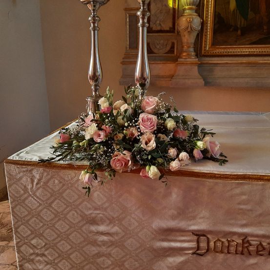 Eine Blumenarrangement mit rosa und weißen Rosen steht auf einem Tisch mit goldenen Kanten. Zwei silberne Kerzenständer stehen hinter den Blumen. Ein Bilderrahmen ist an der Wand befestigt.