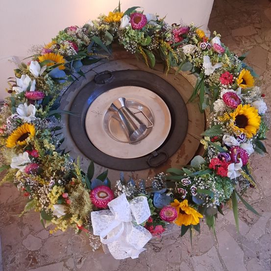 Ein Blumenkranz mit einer Mischung aus weißen, gelben und roten Blumen sowie grünen Blättern liegt auf einem gefliesten Boden. In der Mitte ist eine metallische Platte platziert. Die Platte zeigt ein Kreuzsymbol.