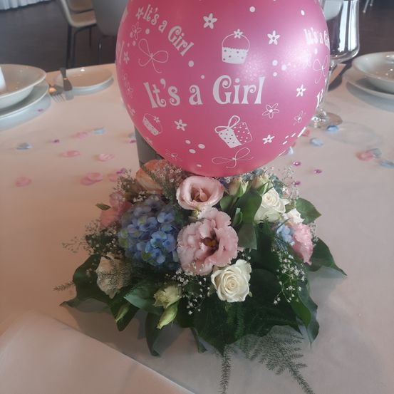 Ein rosafarbener Luftballon mit der Aufschrift Es ist ein Mädchen steht auf einem Blumenarrangement. Es gibt blaue, rosa und weiße Blumen sowie grüne Blätter.