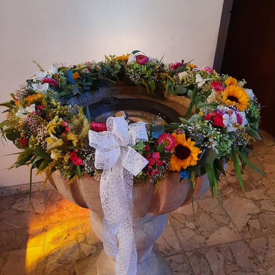 Ein Taufbecken aus Stein, geschmückt mit einer kreisförmigen Anordnung verschiedener Blumen, darunter Sonnenblumen, Rosen und Gänseblümchen, verziert mit einer weißen Schleife.