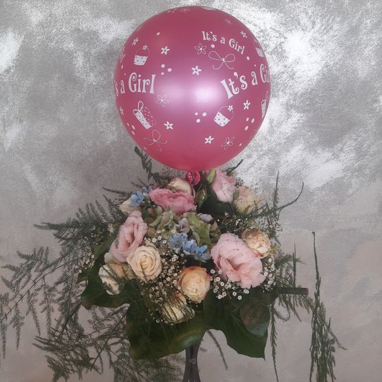 Ein rosafarbener Ballon mit der Aufschrift 'Es ist ein Mädchen' befindet sich über einer Blumenarrangement mit verschiedenen Blumen und Blättern.